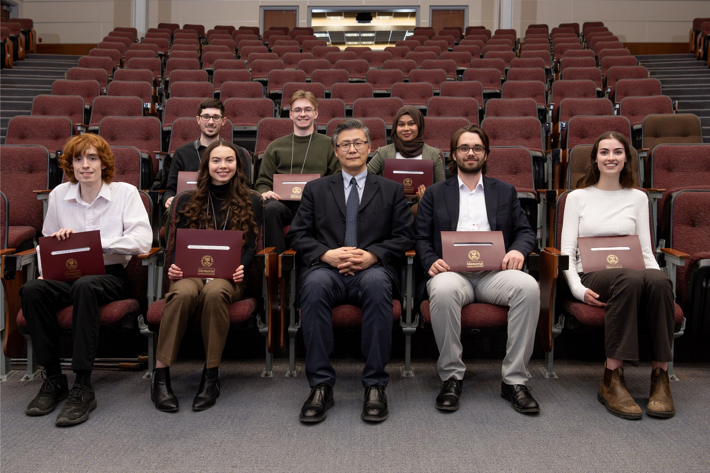 A group of eight poses in an auditorium, with a central figure in a suit and seven young individuals holding MUN certificates.